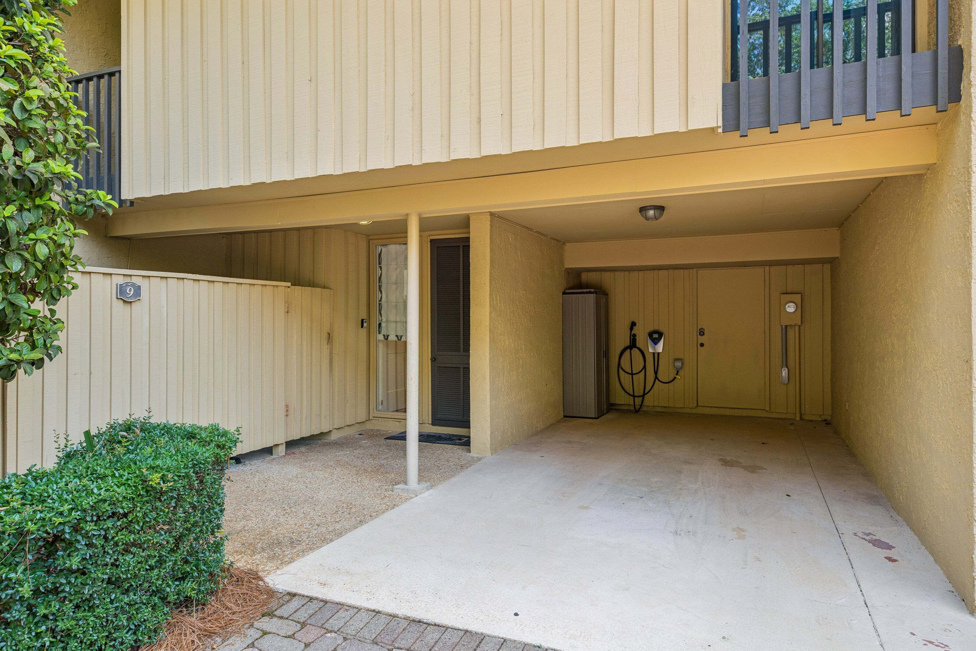 A parking area with a concrete floor, a small green bush, and a building with a beige exterior.