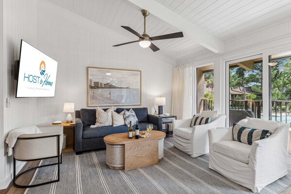 A modern living room with a sofa, armchairs, a coffee table, and a ceiling fan. There is a framed picture on the wall and a sign for 'HOST & HOME Luxury Vacation Rentals.' - 3BR Beachside Villa with Community Pool, Short Walk to the Beach (Hilton Head Island)