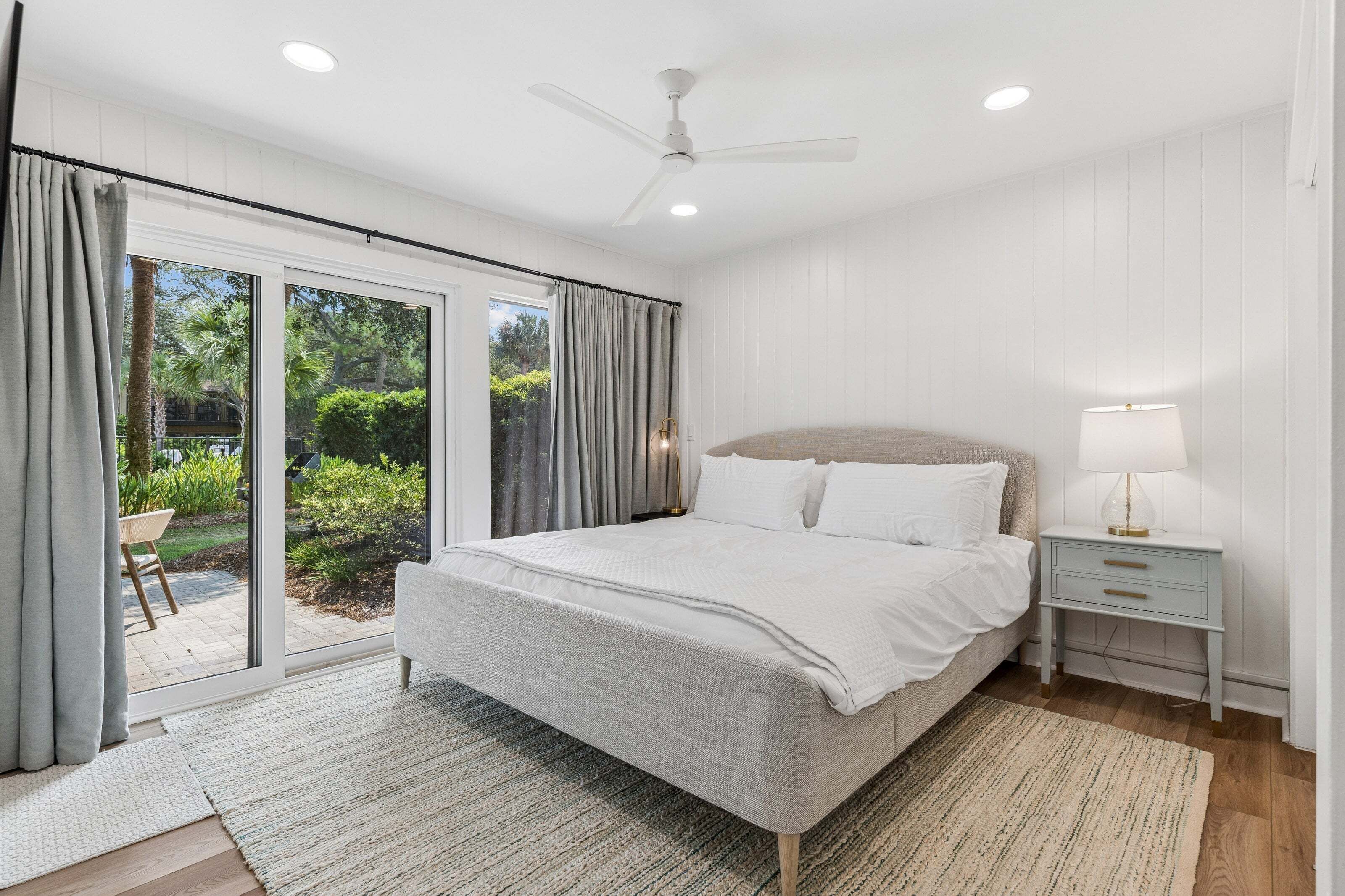 A bedroom with a bed, nightstands, a lamp, a ceiling fan, and a view of a garden through large sliding doors.