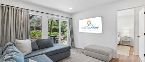 A modern living room with a grey sofa, a grey ottoman, and a large window with curtains. A sign reading 'HOST & Home' is visible on the wall.