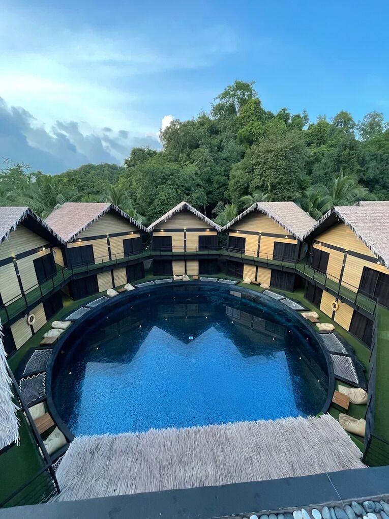Una piscina circular rodeada de bungalows con techo de paja, sobre un exuberante telón de fondo verde.