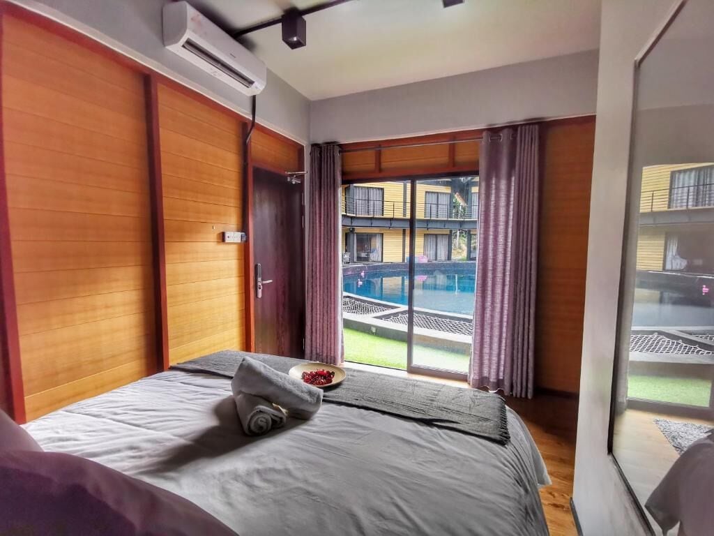 A hotel room with a bed, an air conditioning unit, a window with curtains, and a view of a pool and buildings outside.