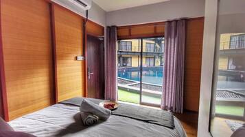 A hotel room with a bed, an air conditioning unit, a window with curtains, and a view of a pool and buildings outside.