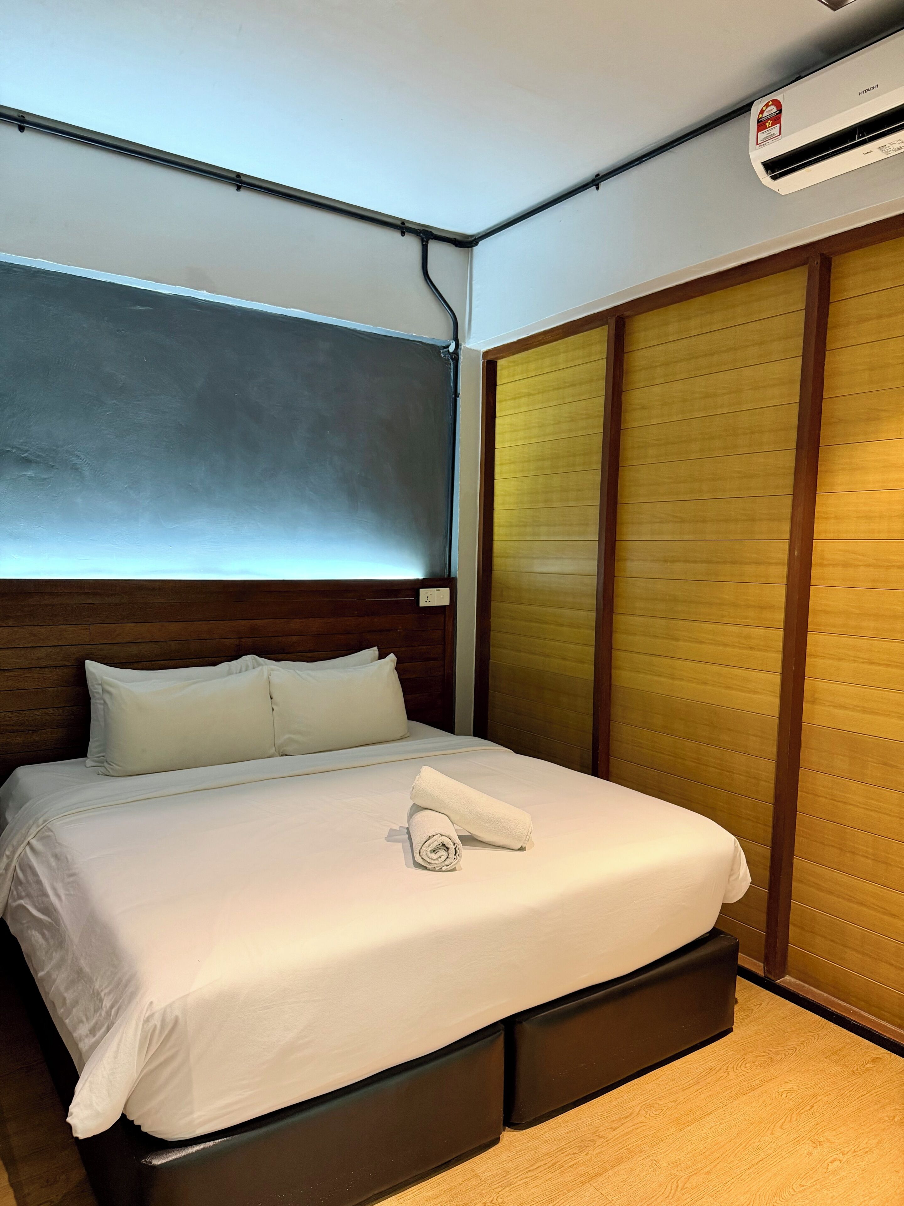 A hotel room with a bed, a wooden headboard, a window with blinds, a wooden wardrobe, and an air conditioning unit.