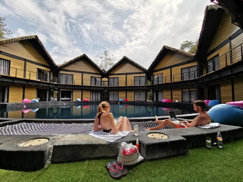 A poolside area with lounge chairs and a netted seating area, surrounded by wooden buildings with a thatched roof.