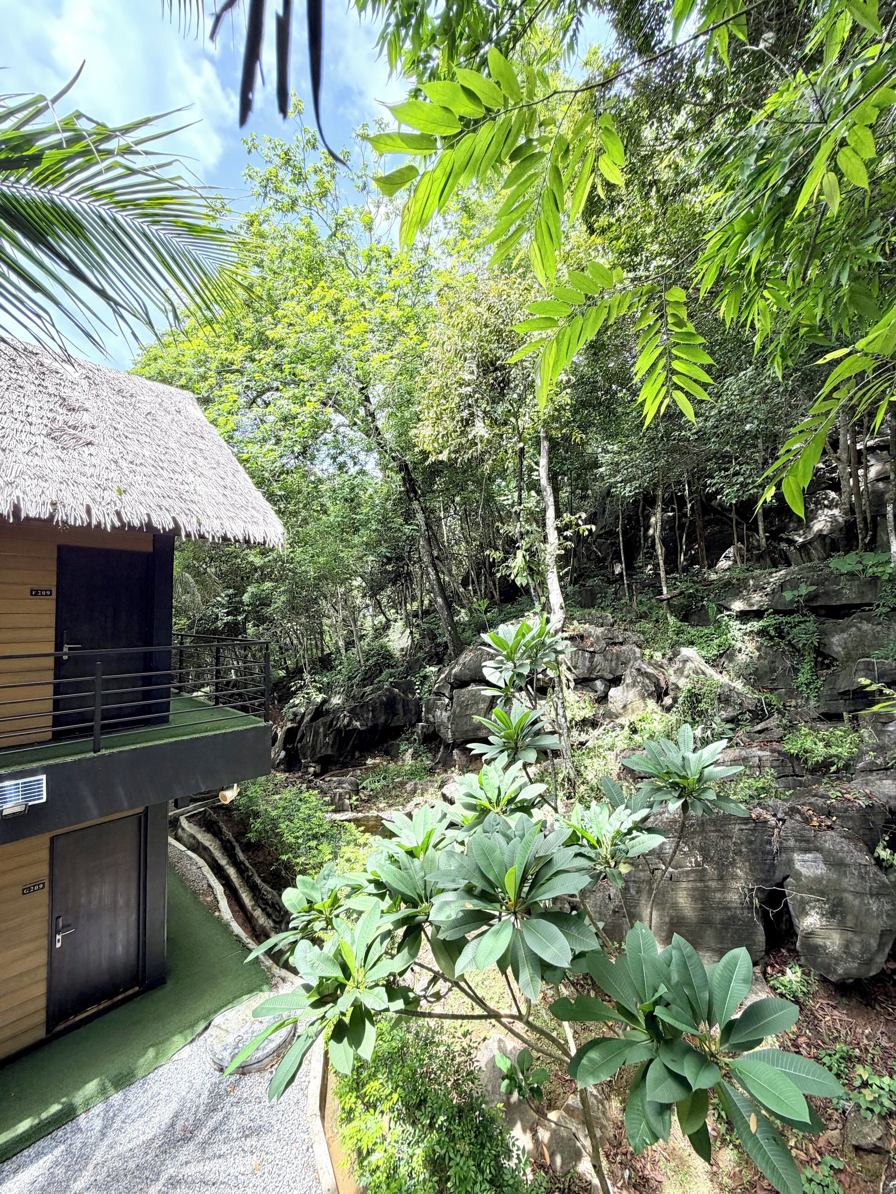 A thatched-roof structure with a balcony, surrounded by lush greenery and rocky terrain.