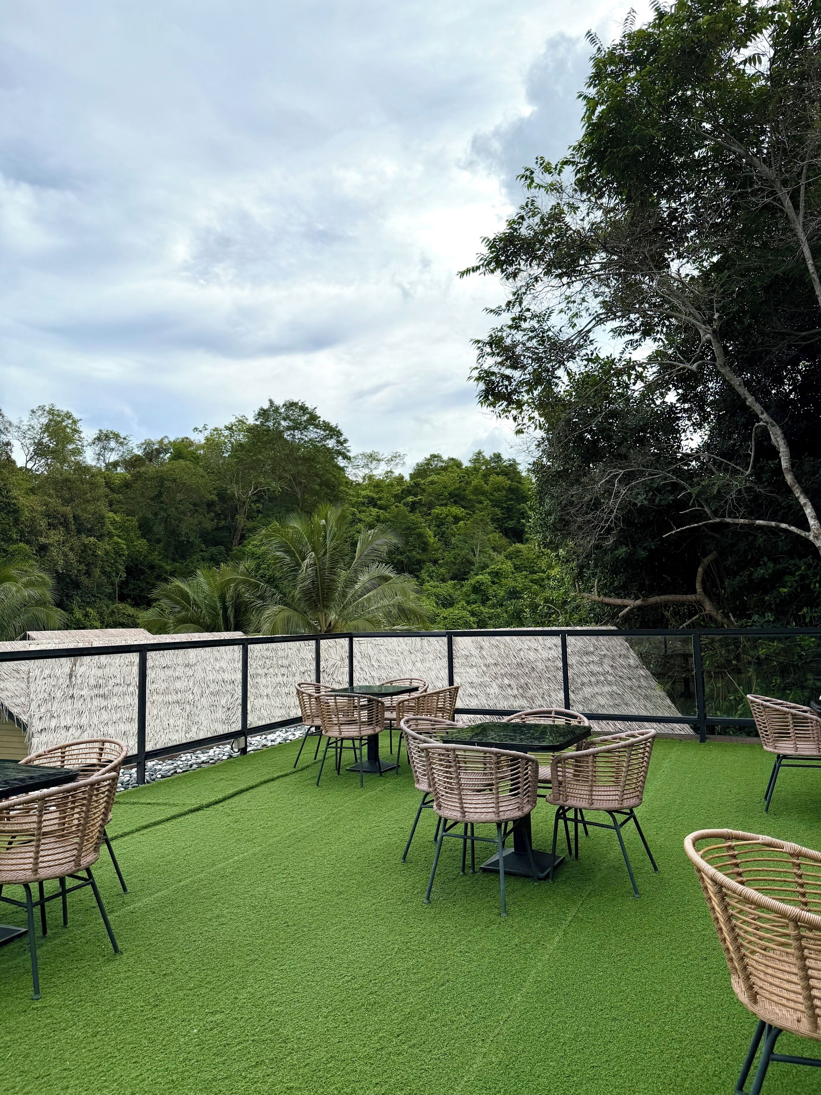 An outdoor seating area with tables and wicker chairs, surrounded by a white wall and lush greenery.