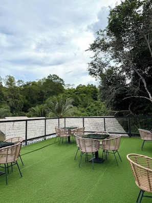Un espacio al aire libre con mesas y sillas de mimbre, rodeado por un muro blanco y abundante vegetación.