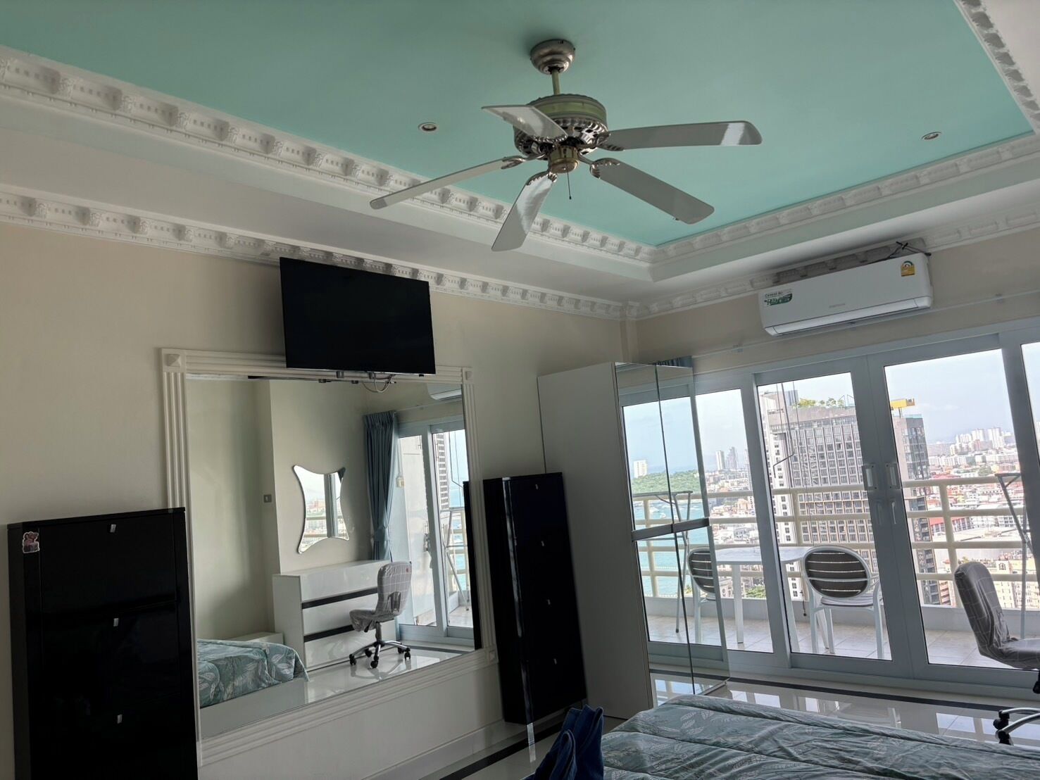 A hotel room with a bed, a desk, a chair, a television, a ceiling fan, and a large window with a city view.