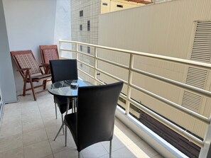 A balcony with a glass table and chairs, a metal railing, and a view of a building with windows and a door.