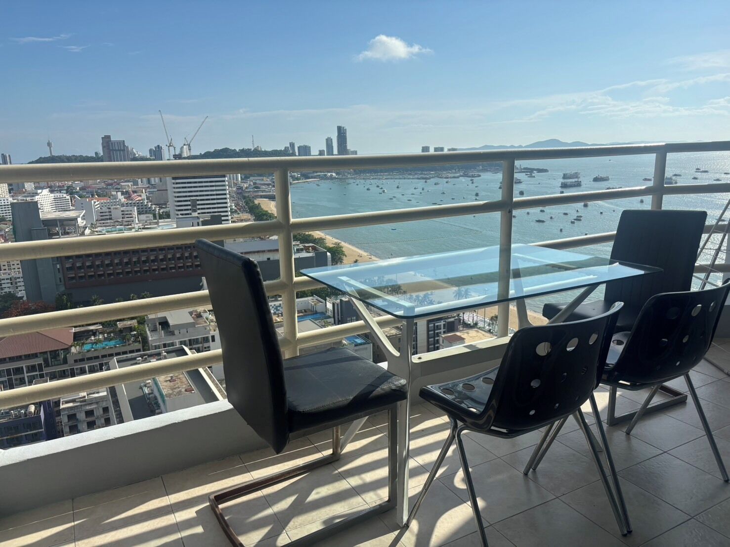 A balcony with a glass table and chairs, overlooking a marina with boats and a cityscape.