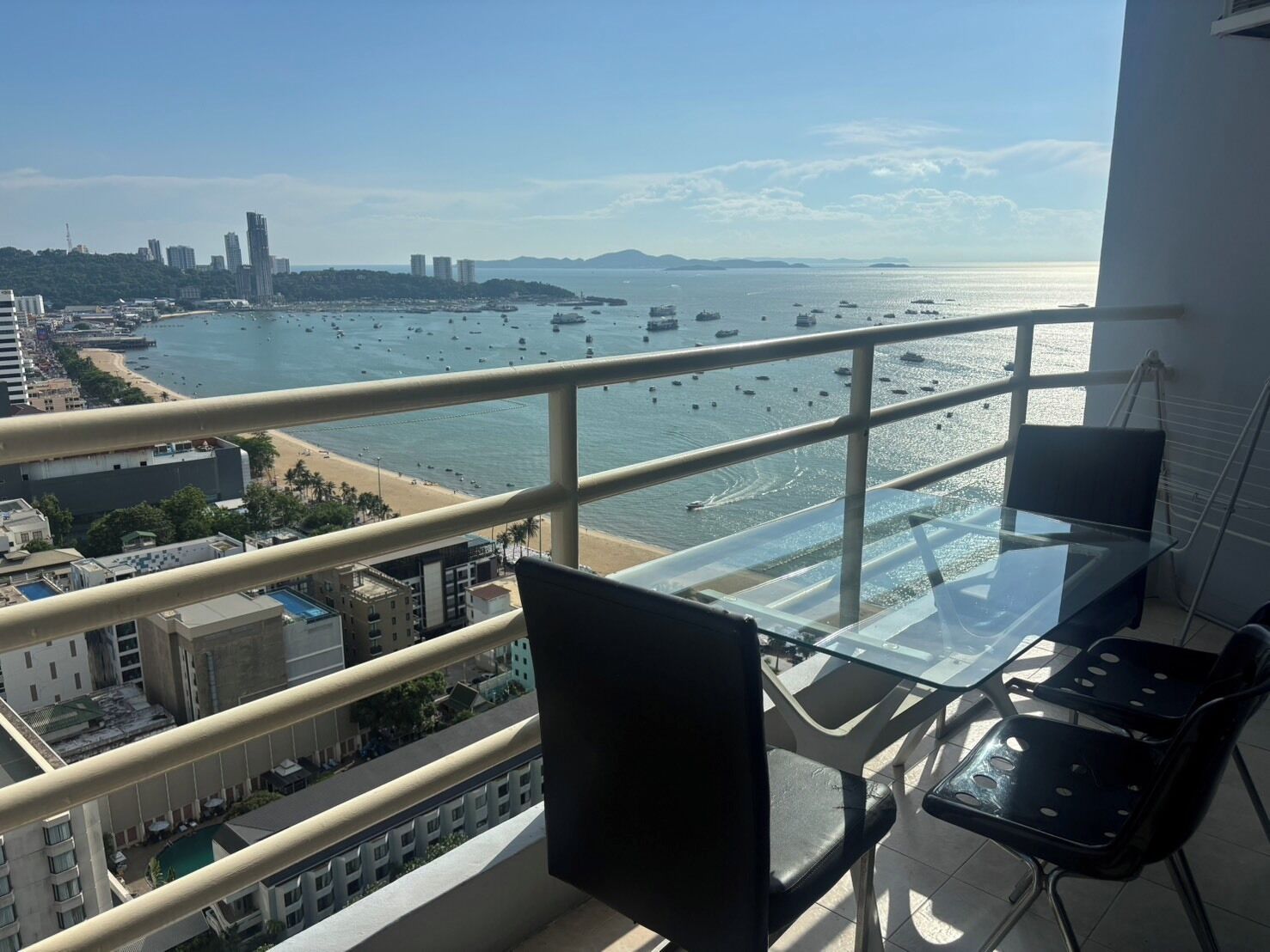 Un balcon avec une table et des chaises en verre, donnant sur une plage où l’on aperçoit des bateaux, des bâtiments et un ciel dégagé.
