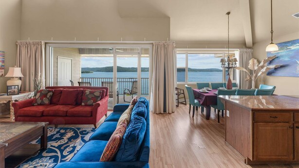 A spacious living area with a red sofa, blue couch, and a dining table set for six. A large window offers a view of the water. - Club Yakov & 3bdr Lakefront (Hollister)