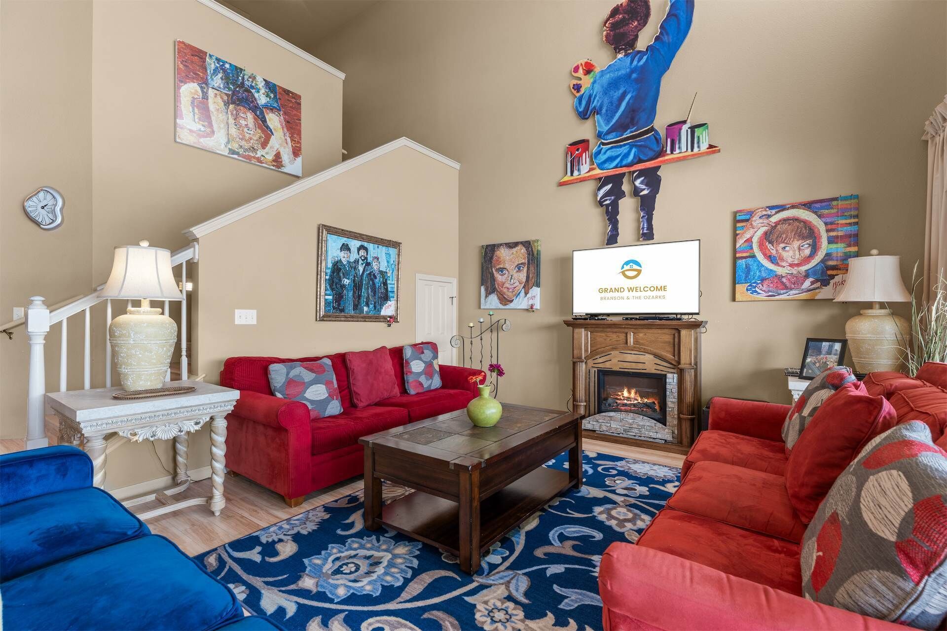 Un salon avec un canapé rouge, un canapé bleu, une table basse en bois et une cheminée. Des tableaux ornent les murs et un téléviseur est fixé au-dessus de la cheminée.