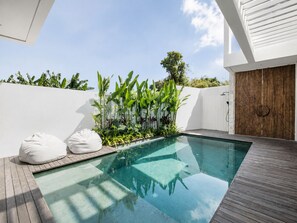A modern outdoor pool area with a wooden deck, a white wall, and a variety of tropical plants. - Casa Rio | 3BR Enclosed Living & Pool (Kuta Utara)