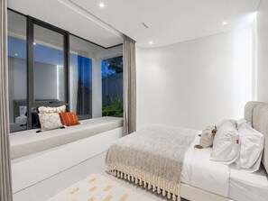 A modern bedroom with a large bed, a window seat, and a view of the outdoors. - Casa Rio | 3BR Enclosed Living & Pool (Kuta Utara)