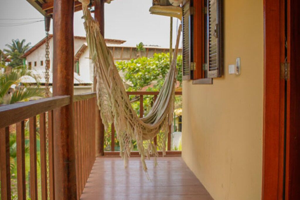Un balcon en bois avec un hamac suspendu, une balustrade en bois et une vue sur la verdure et les bâtiments en arrière-plan.