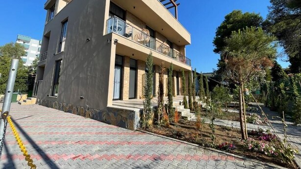 Un bâtiment moderne à deux étages, doté de balcons, d’un espace d’entrée pavé et d’un jardin agrémenté de diverses plantes.