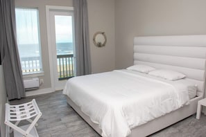 A bedroom with a large bed, a view of the sea through a window, and a white chair. - Casa Dora Summer Waves (Bolivar Peninsula)