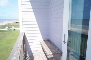 A corner of a room with white walls, a wooden bench, and a glass door leading to a balcony overlooking a beach.