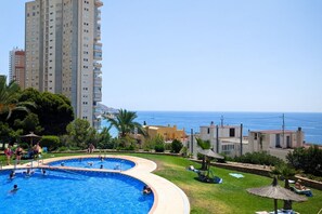 A swimming pool with a curved design, surrounded by a grassy area with lounge chairs and umbrellas, and a tall building in the background. - PINTOR SOROLLA elegant apartment (Benidorm)
