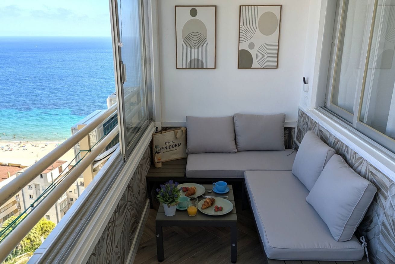 Ein Balkon mit Blick auf das Meer, eine Sitzecke mit Sofa, ein kleiner Tisch mit Frühstücksgebäck und zwei abstrakte Wandbilder.