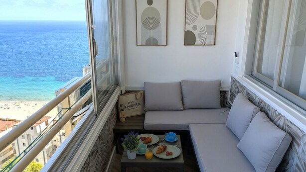 A balcony with a view of the ocean, a sofa, a small table with breakfast items, and two abstract wall art pieces. - PINTOR SOROLLA elegant apartment (Benidorm)