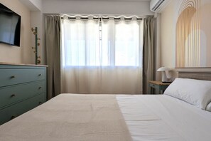 Ein Hotelzimmer mit Bett, Wandfernseher, Schreibtisch mit Lampe und einem fensterbepflanzten Fenster.