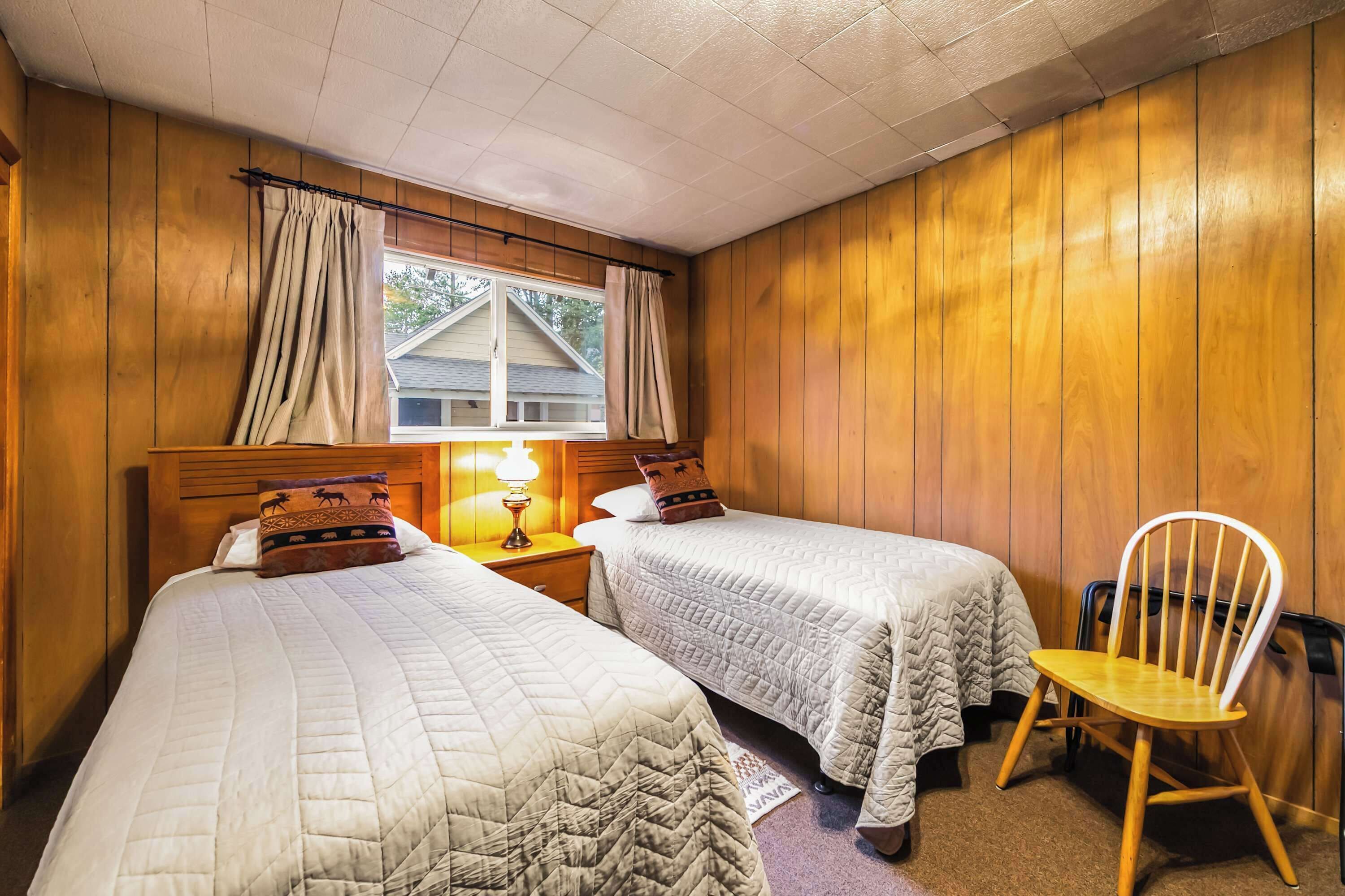 Een kamer met twee bedden, een nachtkastje met lamp, een stoel en houten wanden.