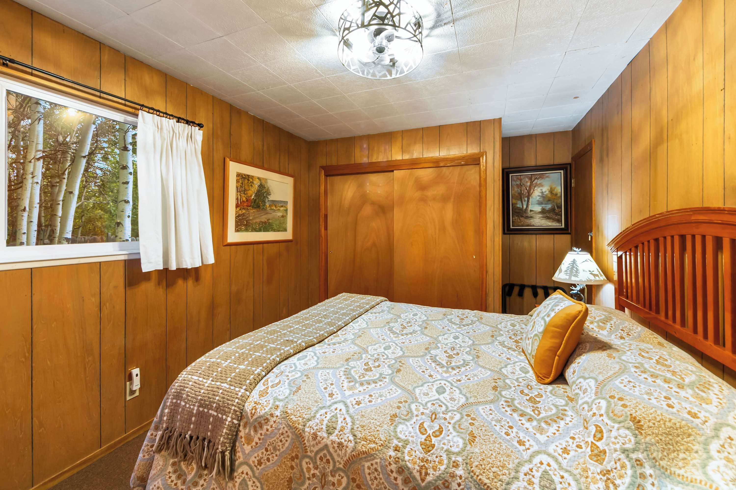 Een slaapkamer met houten lambrisering, een bed met beddengoed met een patroon, een raam met gordijnen en een plafondventilator.