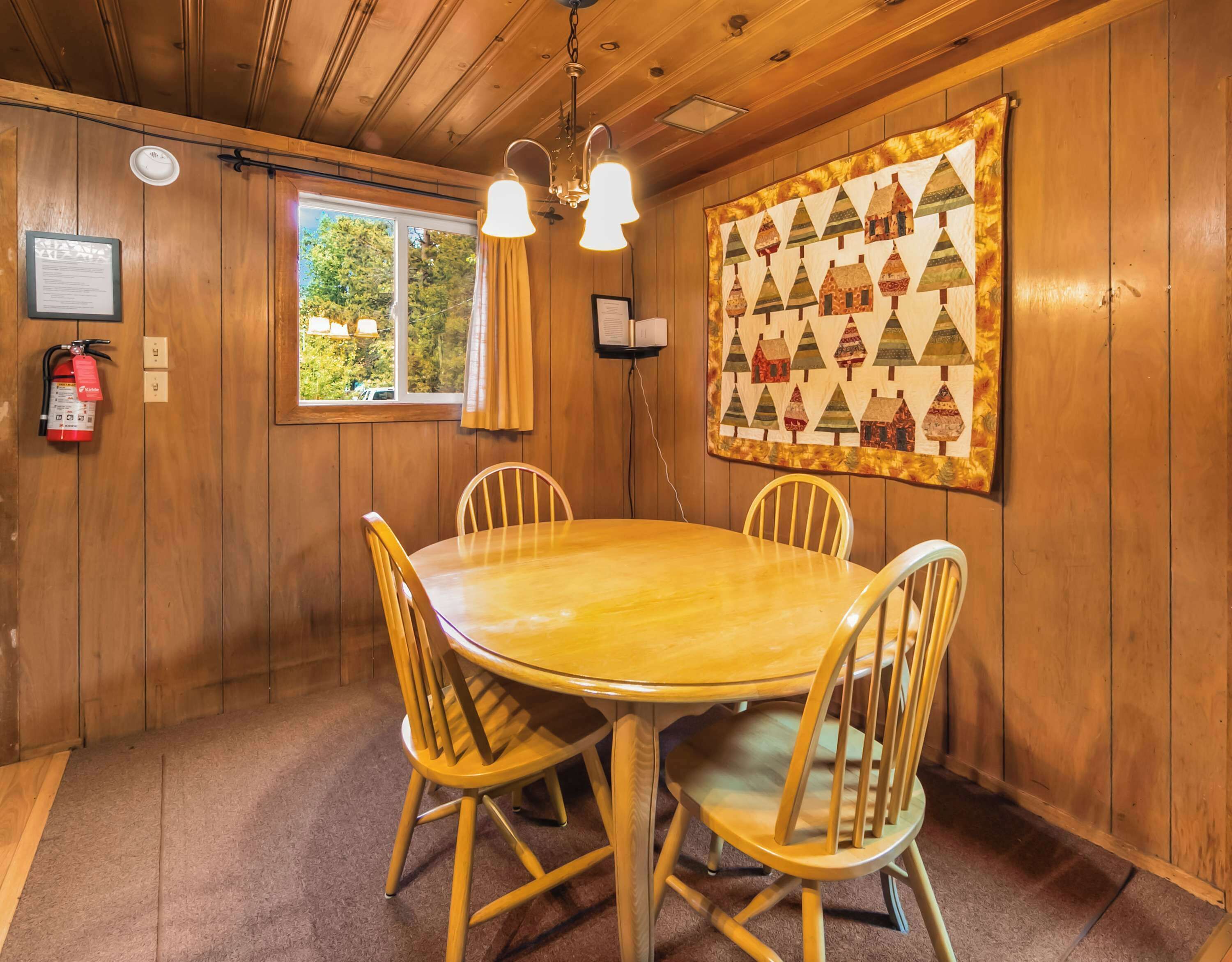 A wooden-paneled room with a round table and chairs, a fire extinguisher, a window with curtains, and a quilted wall hanging.
