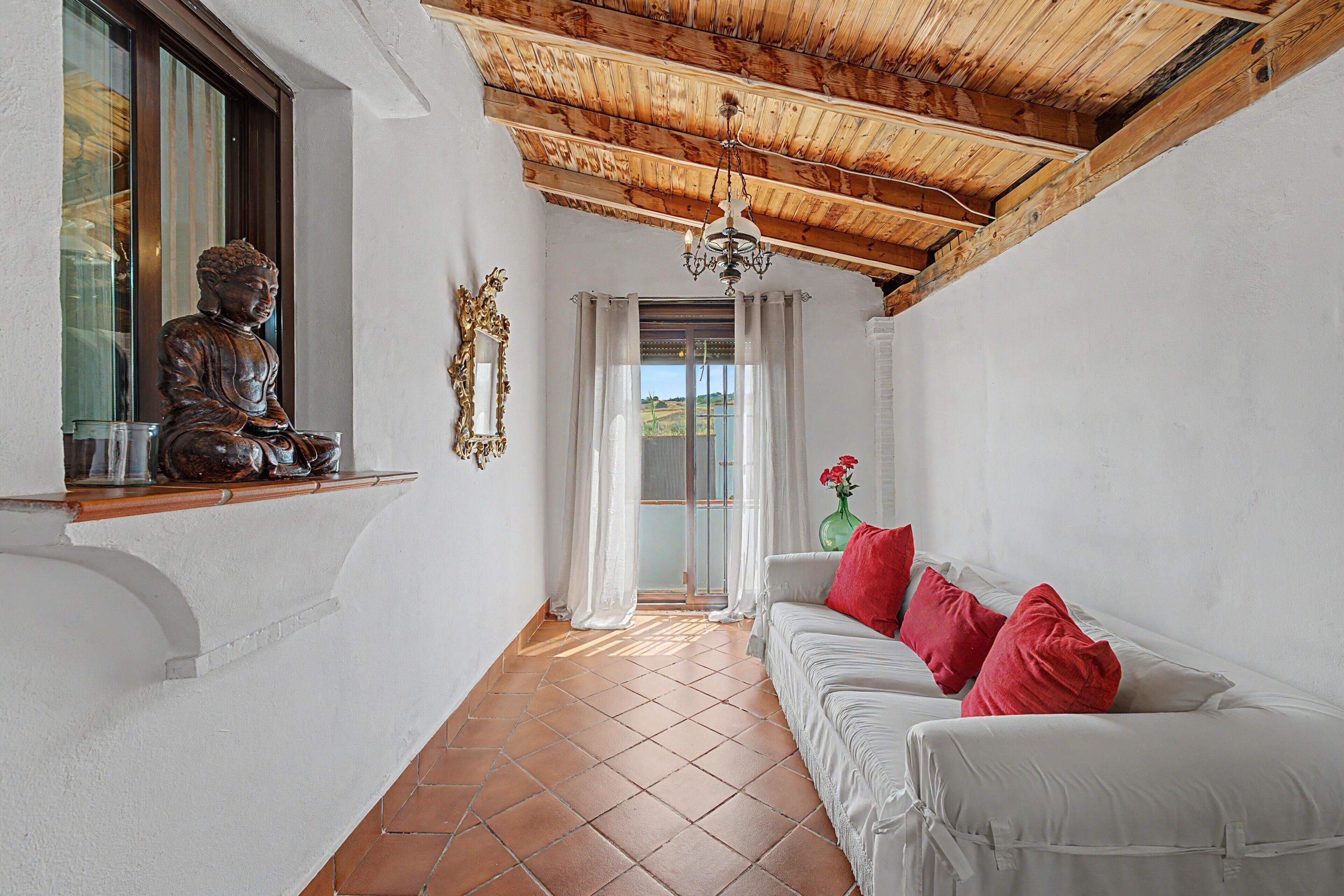 Ein Zimmer mit Buddha-Statue, einem weißen Sofa mit roten Kissen, einer Holzbalkendecke und Blick durch ein Fenster.