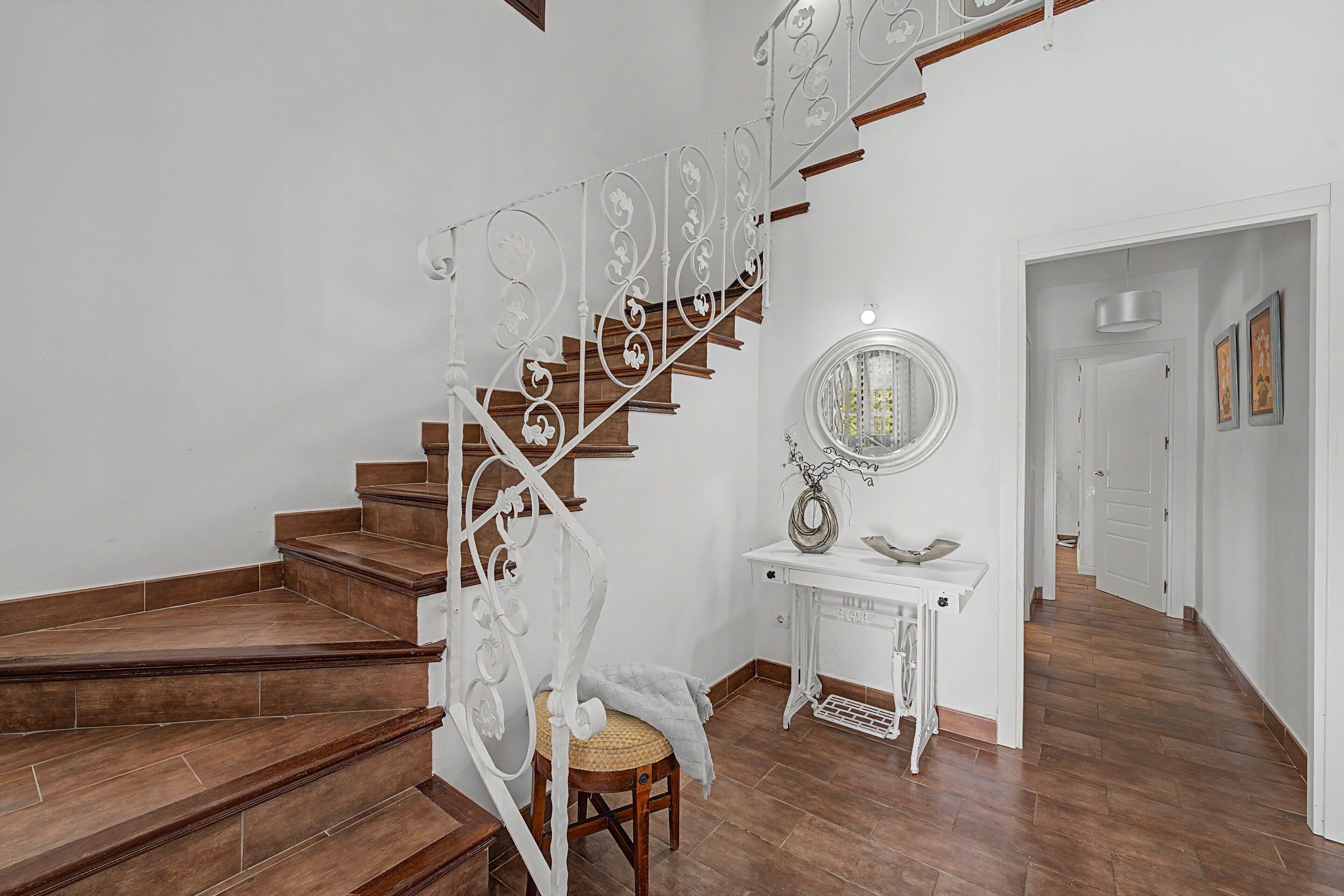Una escalera con barandilla metálica blanca, suelo de madera, una consola blanca con espejo y una puerta que da a otra habitación.