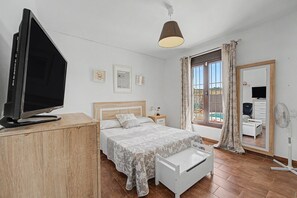 Um quarto com uma cama, uma televisão sobre um móvel de madeira, uma mesa de cabeceira, uma lâmpada, uma janela com cortinas e vista para o exterior.