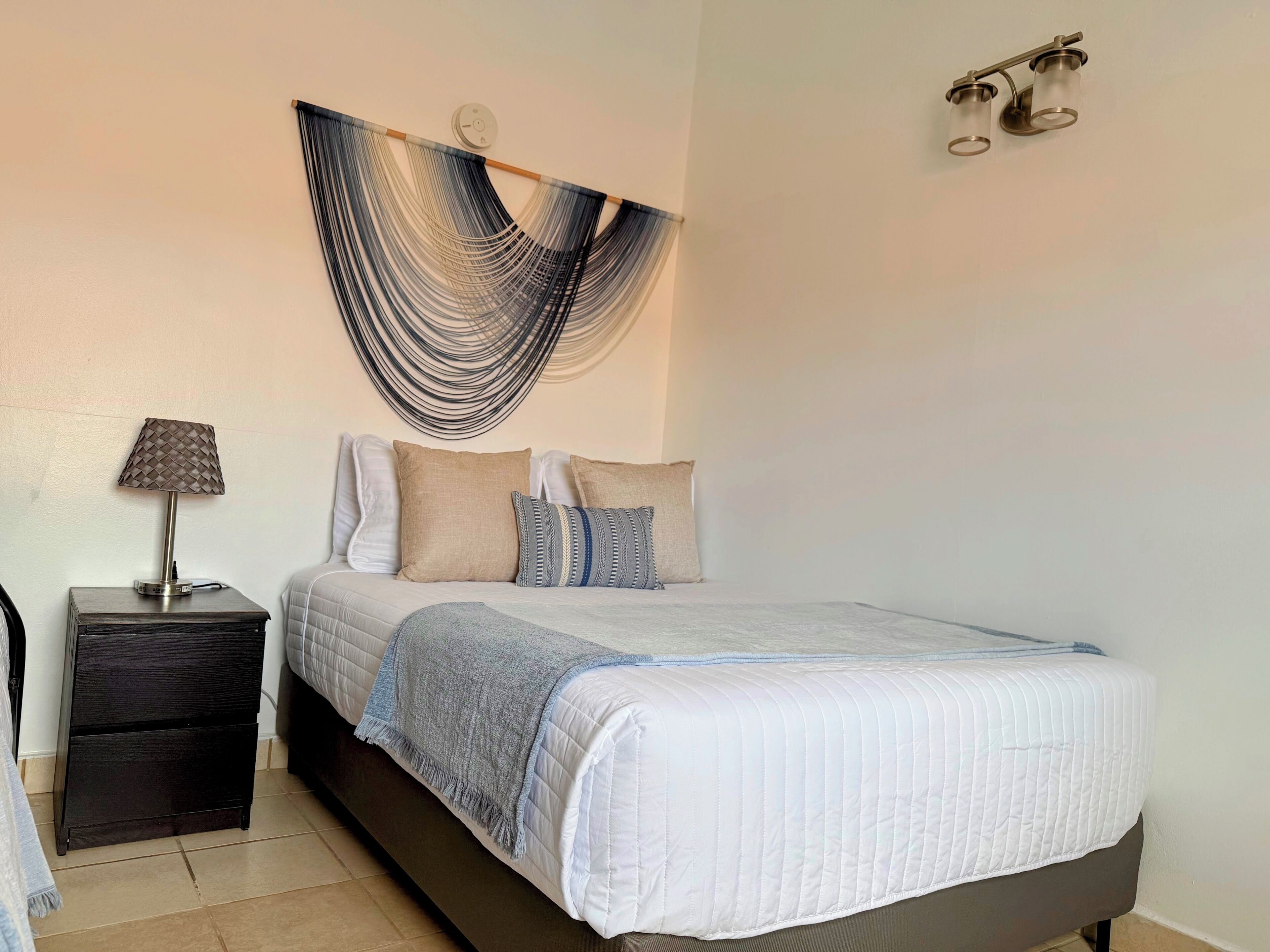 A bedroom with a bed, bedside table, lamp, wall art, and a wall-mounted light fixture.