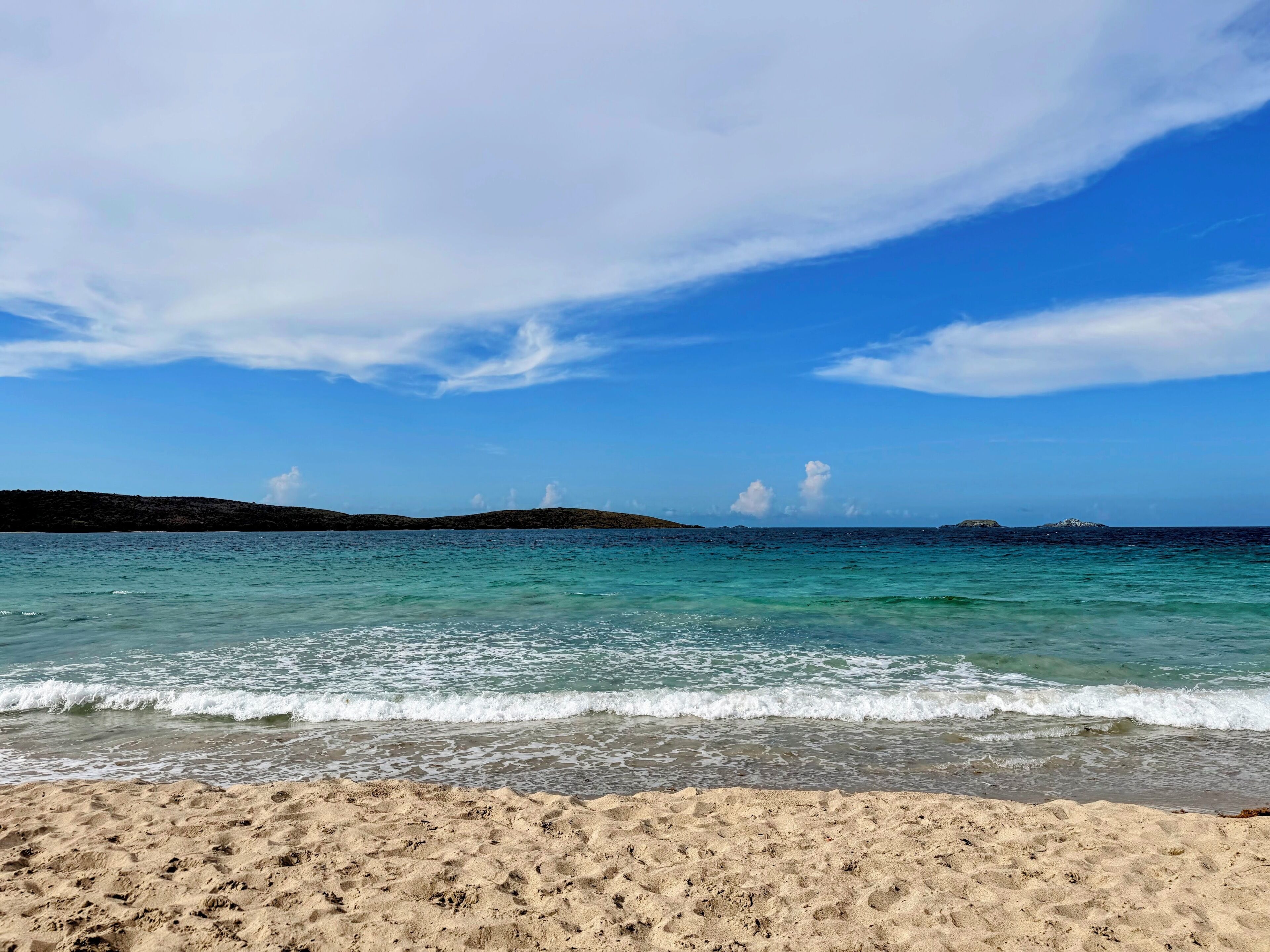 Une plage à l’eau turquoise et limpide, aux vagues douces et bordée de collines lointaines.
