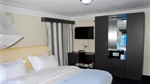 A hotel room with a bed, a desk, a chair, a wardrobe, a television, and a clock on the wall. - KAA HIGHWAY HOTEL (Mukono)