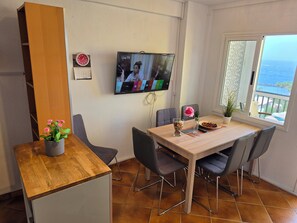 Ein Essbereich mit einem Holztisch, grauen Stühlen, einer Topfpflanze und einem an der Wand befestigten Fernseher.