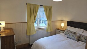 Et soveværelse med seng, sengeborde, et vindue med gardiner, en radiator og et trænatbord.