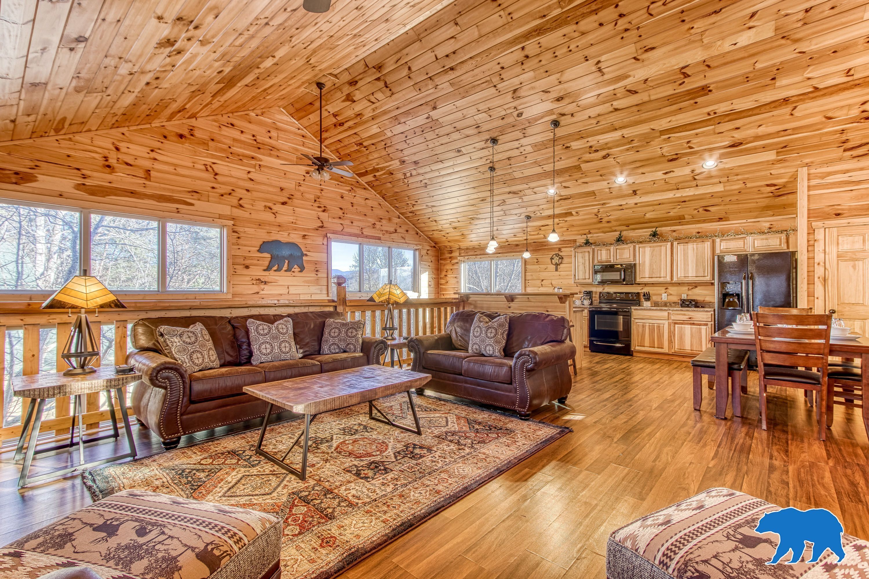 A spacious cabin interior with a wooden ceiling, a cozy living area with a sofa and coffee table, a kitchen with modern appliances, and large windows offering views of the surrounding forest.
