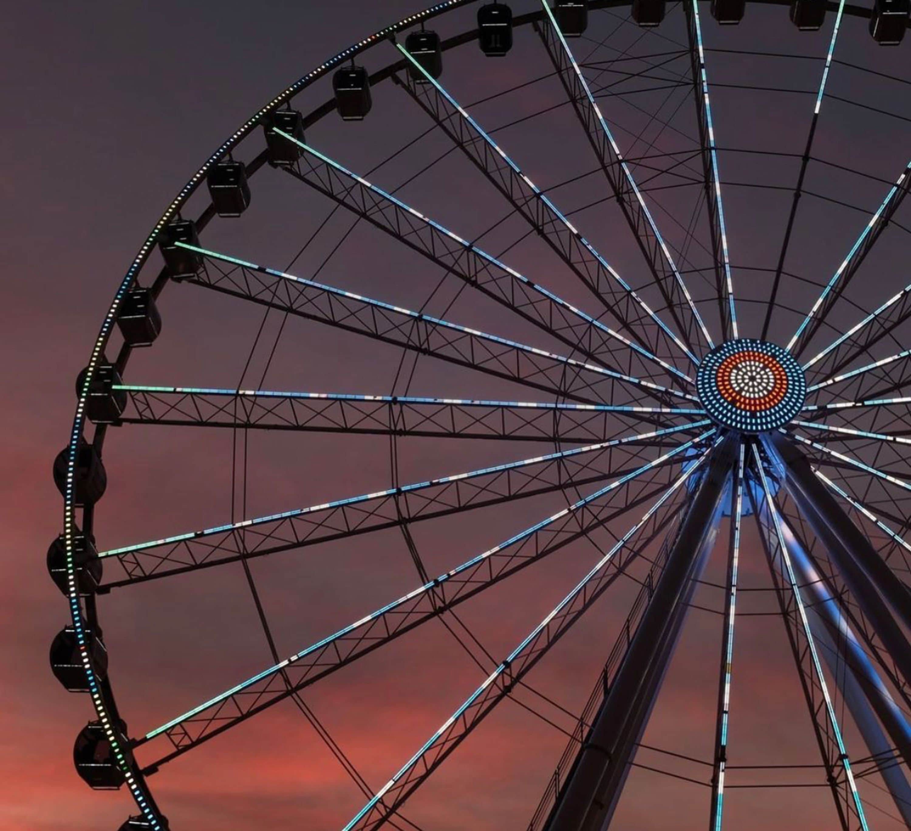 Ein großes Riesenrad mit einer zentralen Achse und mehreren Gondeln, vor einem Sonnenuntergangshimmel.