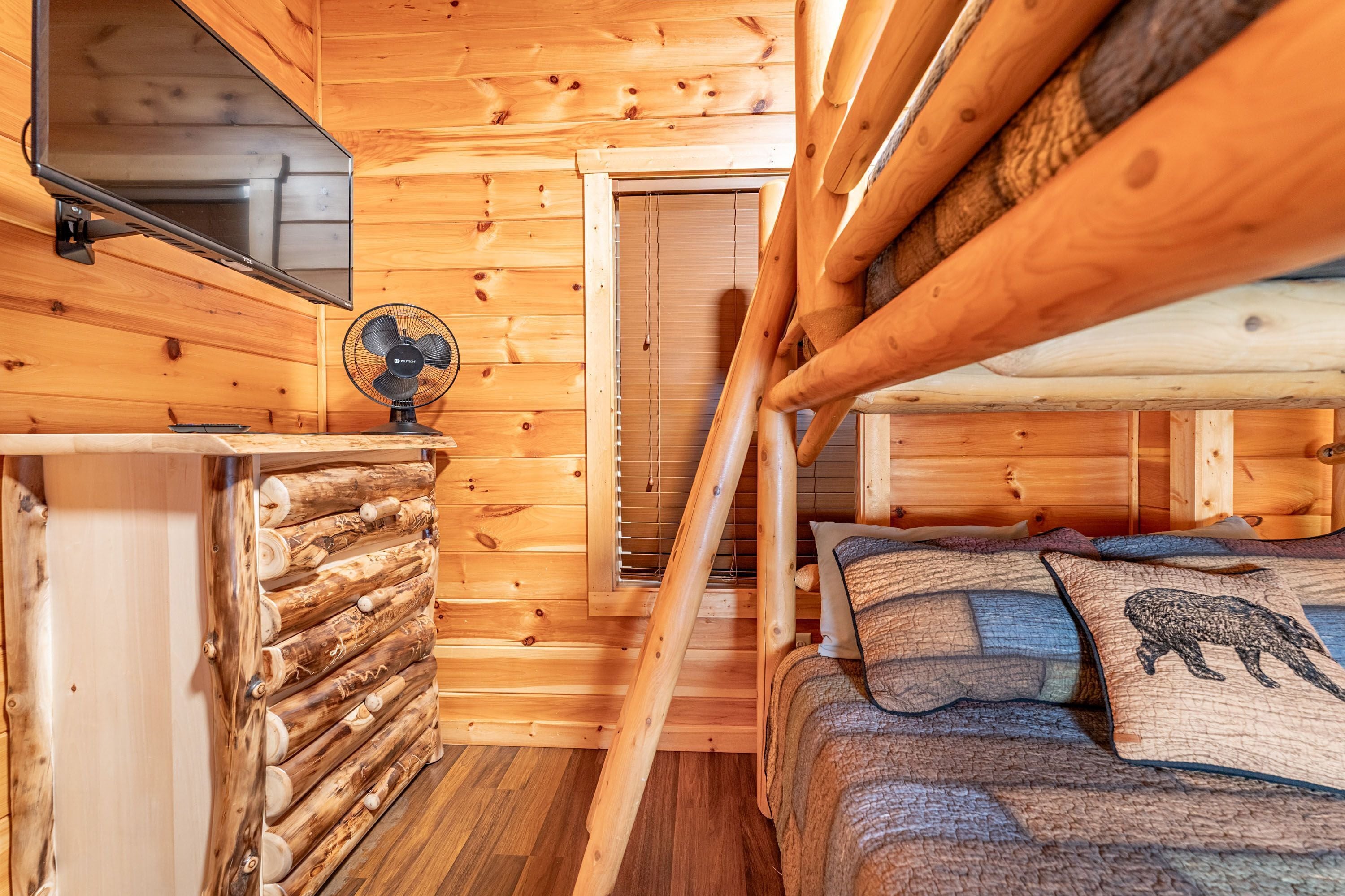 Interior de uma cabana de madeira com uma cama beliche, uma escrivaninha pequena, um ventilador e uma televisão.