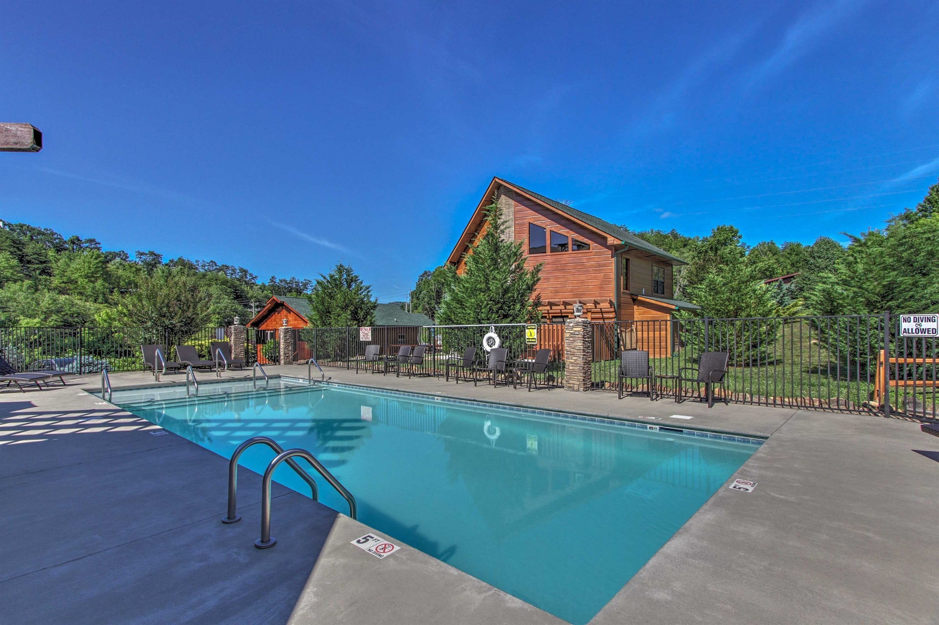 A clear swimming pool with a lifeguard chair, surrounded by lounge chairs and a wooden building with a stone facade.