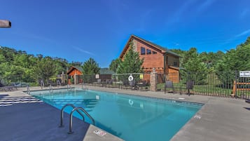 A clear swimming pool with a lifeguard chair, surrounded by lounge chairs and a wooden building with a stone facade.