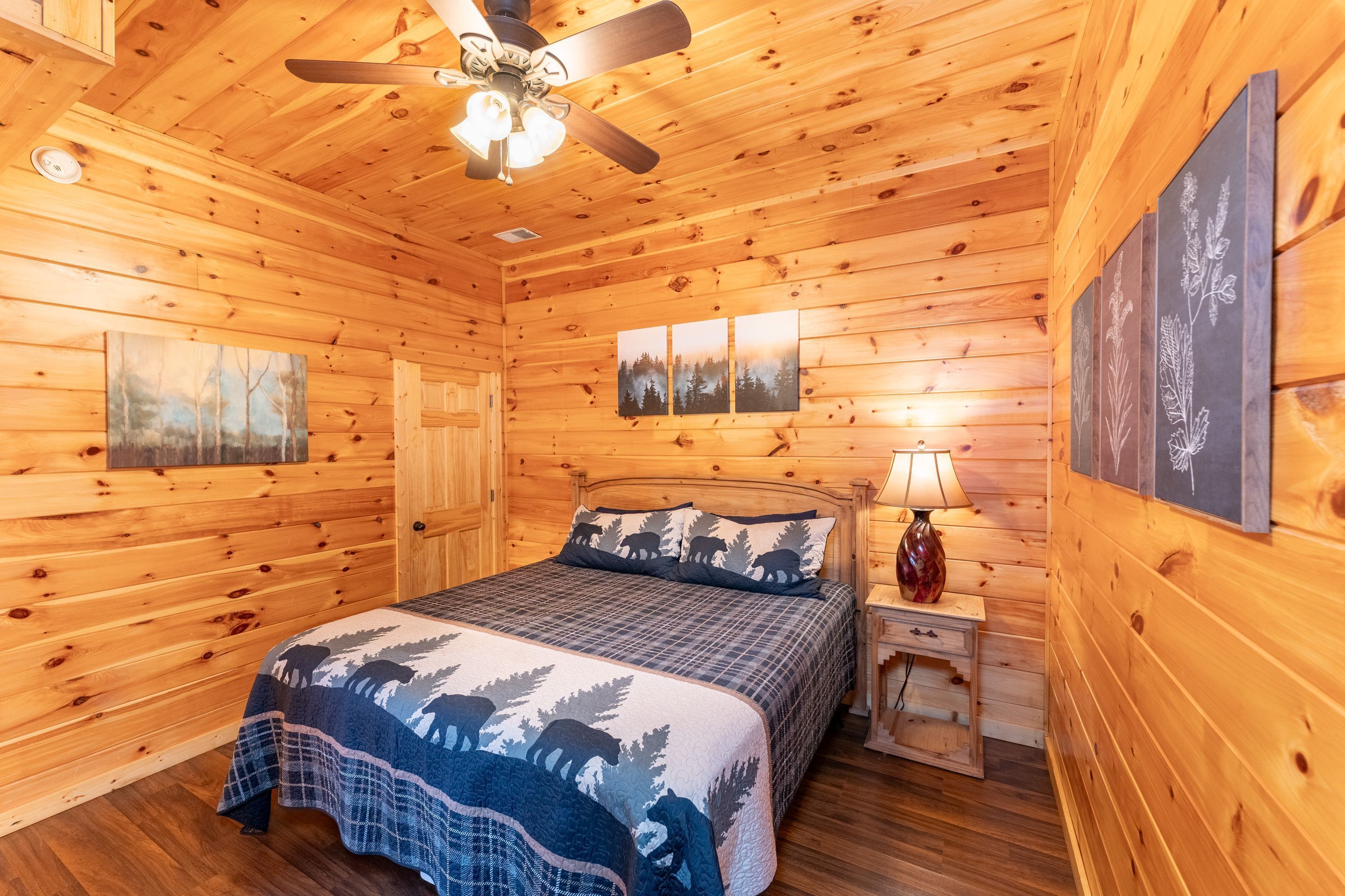 A wooden cabin bedroom with a bed, a nightstand, a lamp, a ceiling fan, and wall-mounted artwork.