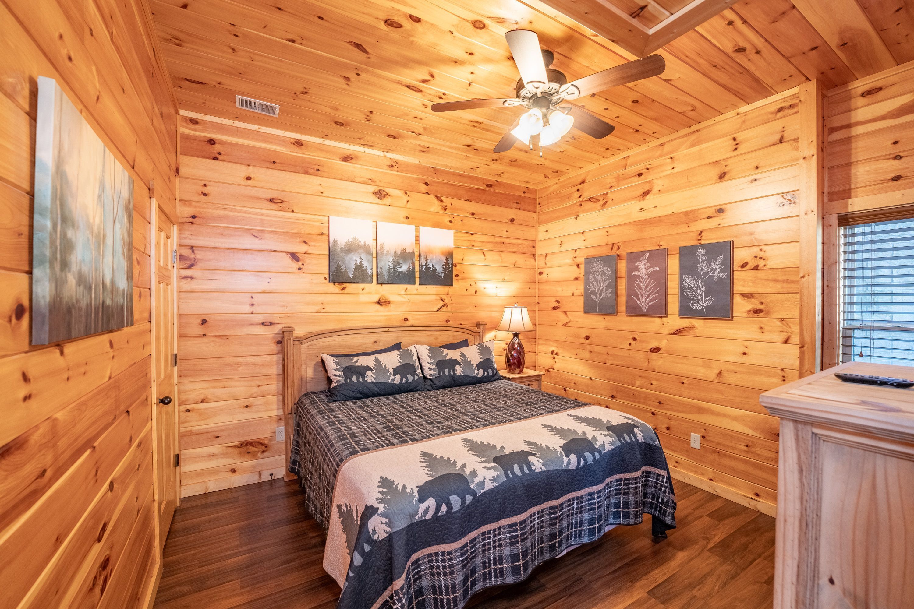 A wooden cabin bedroom with a bed, a ceiling fan, and wall-mounted artwork.