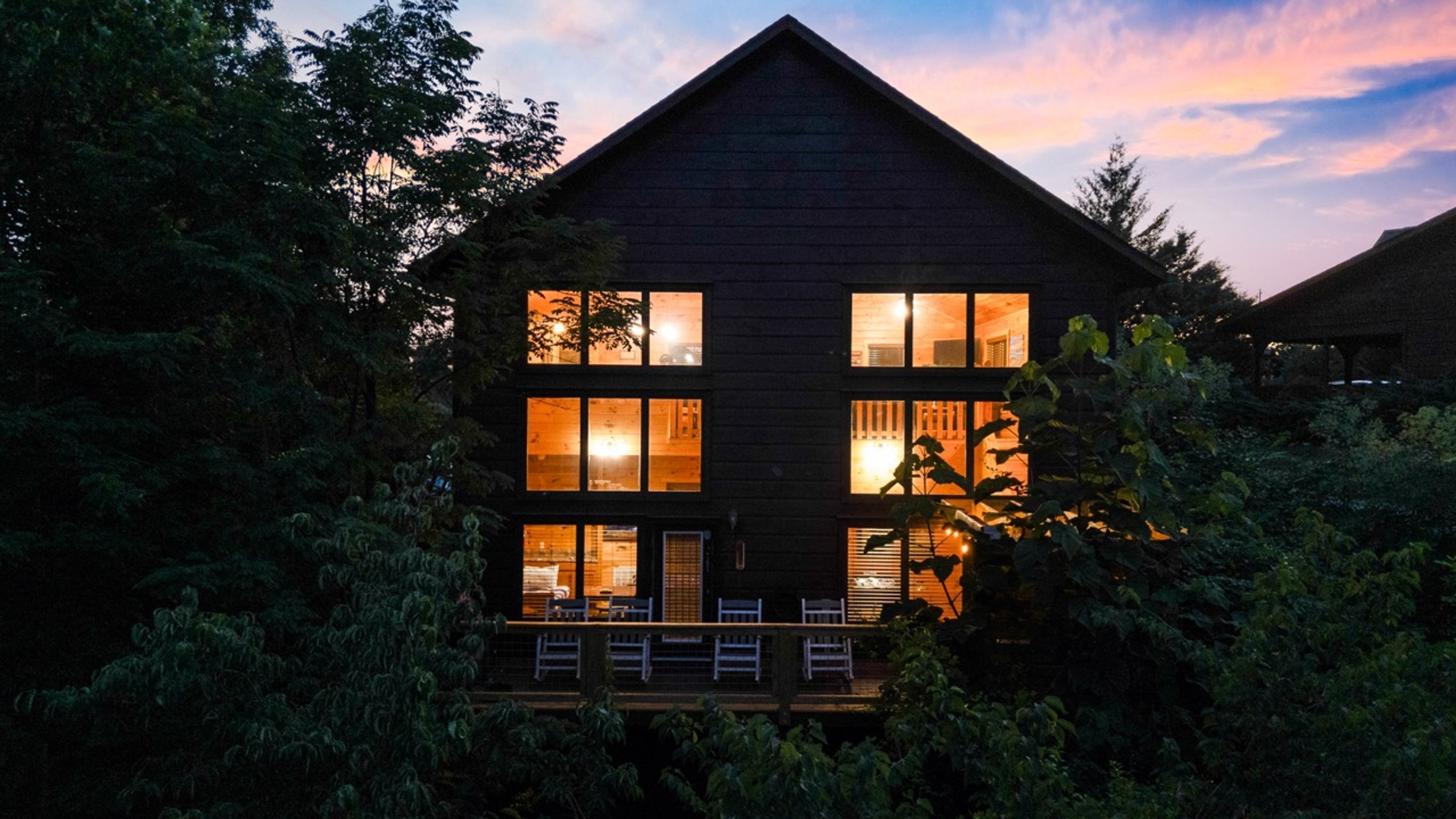 Une maison en bois à deux étages, dotée de nombreuses fenêtres et entourée d’un feuillage dense.