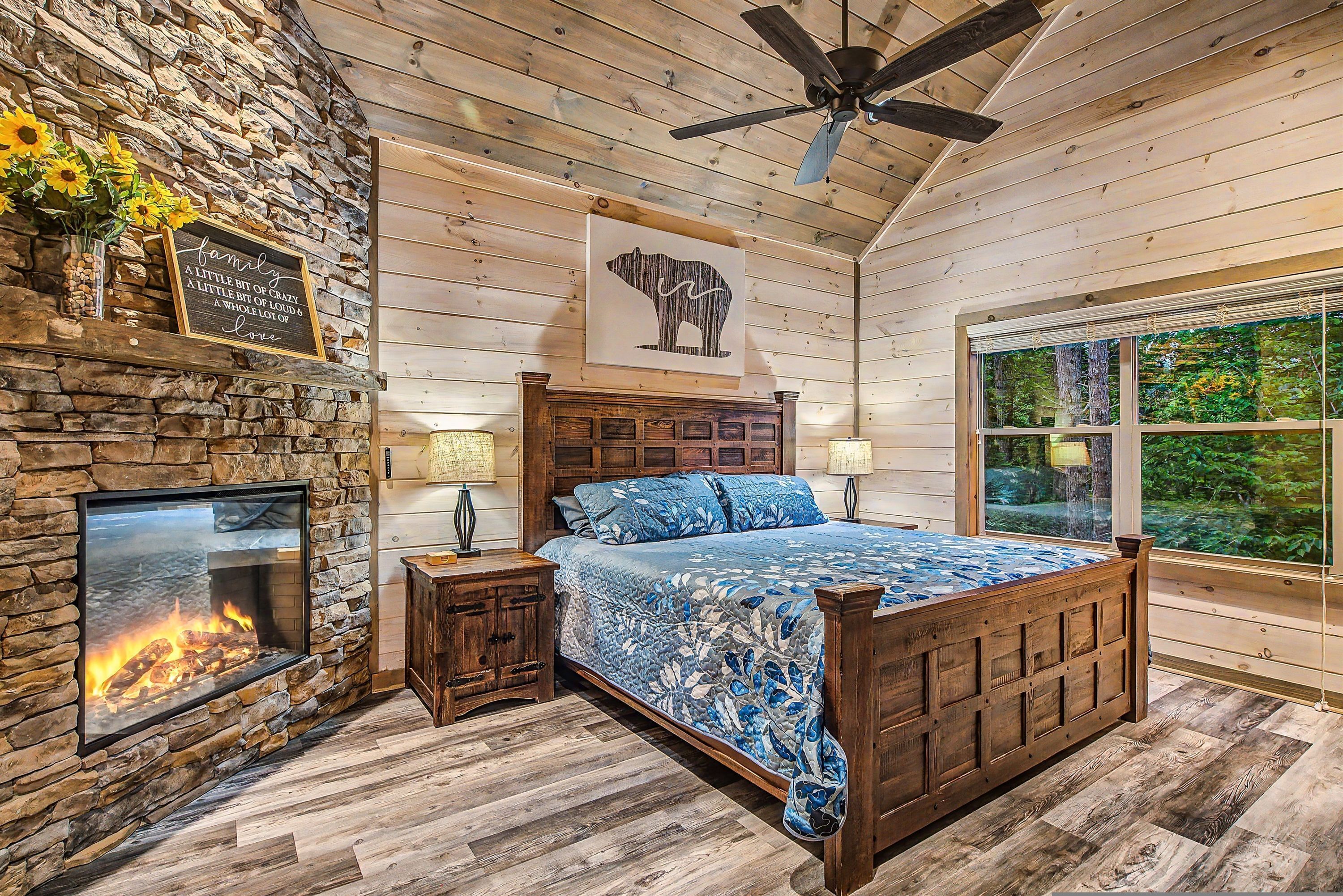 A bedroom with a stone fireplace, a bed with a blue floral bedspread, a wooden headboard, and a large window with a view of greenery.