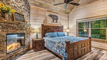 Ein Schlafzimmer mit Kamin aus Stein, einem Bett mit blauem Blumenüberwurf, einem Holz-Kopfteil und einem großen Fenster mit Blick auf Grünflächen.
