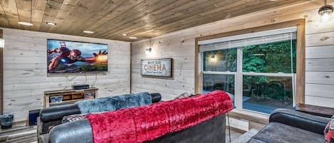 A cozy living room with a large flat-screen TV, a cinema sign, and a large window with a view of greenery.